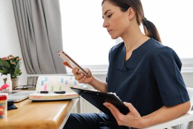 Evde masa başında çalışırken cep telefonu kullanan odaklı kadın doktor.