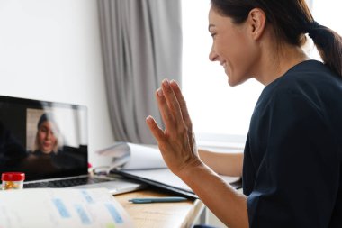 Medikal takım elbiseli mutlu bir kadın dizüstü bilgisayarında video bağlantısıyla konuşuyor ve parlak bir odada masada otururken kamerasına eliyle el sallıyor.