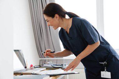 Gülümseyen bir doktor dizüstü bilgisayarın önünde duruyor ve parlak bir odada video konuşmalarıyla konuşuyor.