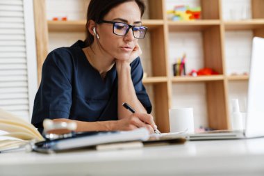 Bir kadın doktor dizüstü bilgisayarın önünde oturuyor kulaklıklar ve gözlüklerle ve parlak bir odadaki deftere bir şeyler yazıyor.