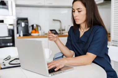 Odaklanmış kadın doktor evde otururken kredi kartı ve laptop kullanıyor.