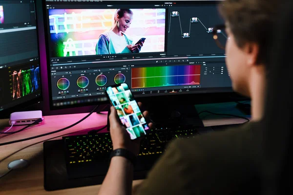 Back view of a young white man videographer working on a project on computer sitting at the desk indoors looking at mobile phone screen