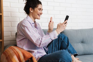 Elinde bir telefon tutan ve odada kanepede otururken kamerasına el sallayan gülümseyen kadın.