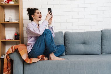 Bacakları kanepede oturan ve ışık odasında telefona bakan kıvırcık, mutlu kadın.