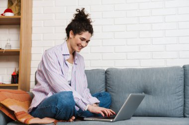 Bacakları kanepede oturan ve ışık odasında dizüstü bilgisayarda yazan mutlu kadının yan görüntüsü.