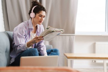 Kulaklıklı kadın kanepede, parlak bir odada otururken dergi okuyor.