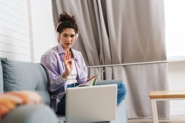 Kulaklıkla dizüstü bilgisayarın önünde oturan ve parlak bir odada kamerasına el sallayan kadın.