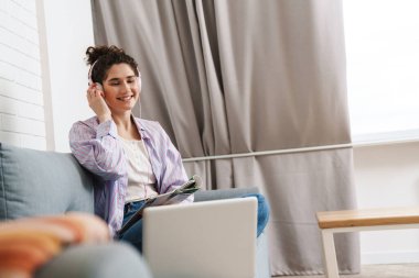 Parlak bir odada dizüstü bilgisayarın önünde oturmuş kulaklıkla müzik dinleyen kıvırcık kadın.