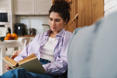 Parlak bir odada kanepede otururken kitap okuyan kadın.