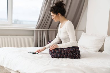 Parlak bir odada ayakları yatakta otururken telefona bakan kadın.