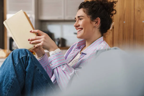 明るい部屋でソファに座っている間に本を読んでいる笑顔の女性の側面図 — ストック写真