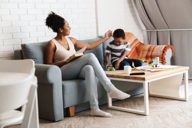 Oğlu evde oyuncaklarla oynarken siyah kadın kitap okuyor.