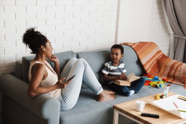 Oğlu evde kitap okurken gülümseyen siyah bir kadın cep telefonu kullanıyor.