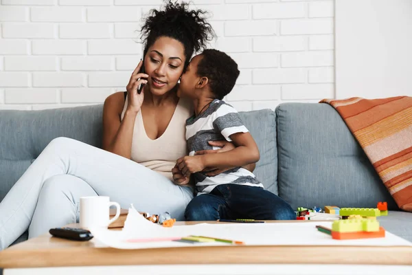 Black smiling woman talking on cellphone while hugging her son at home