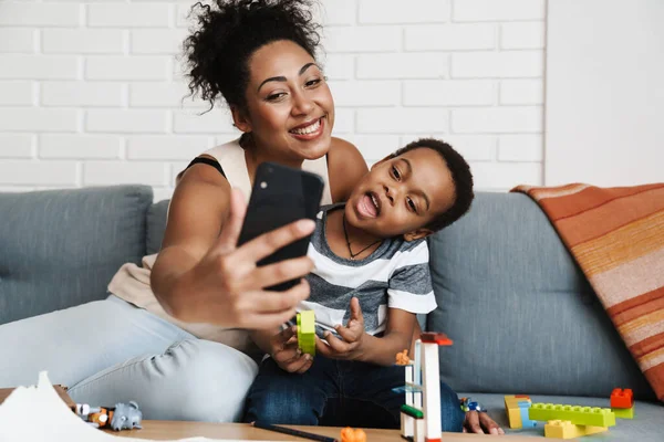Black smiling woman taking selfie photo on cellphone while hugging her son at home