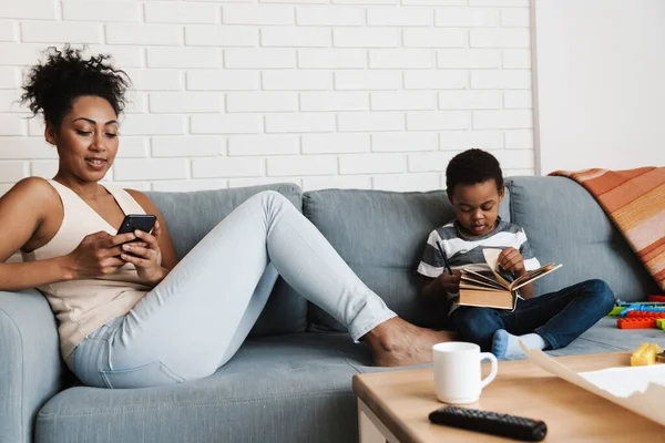 Negro Sonriente Mujer Usando Teléfono Celular Mientras Hijo Leer Libro — Foto de Stock
