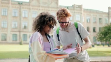 Güneş gözlüğü takan öğrenciler kampüsün dışında kitap izlerken konuşuyorlar.