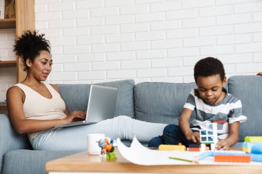 Oğlu evde oyuncaklarla oynarken, siyah kadın dizüstü bilgisayarla çalışıyor.