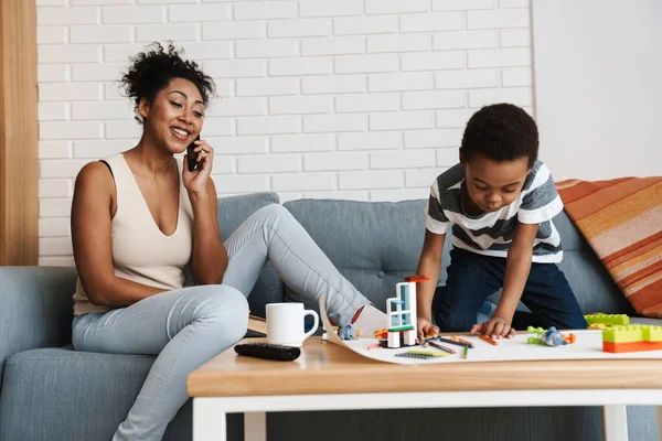 Black smiling woman talking on cellphone while her son playing with toys at home