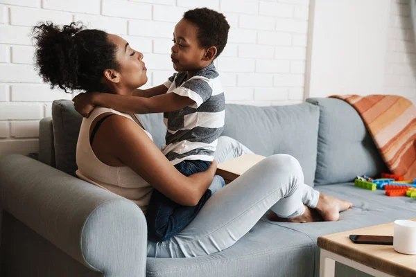Black happy mother and son hugging and making fun while sitting on sofa at home