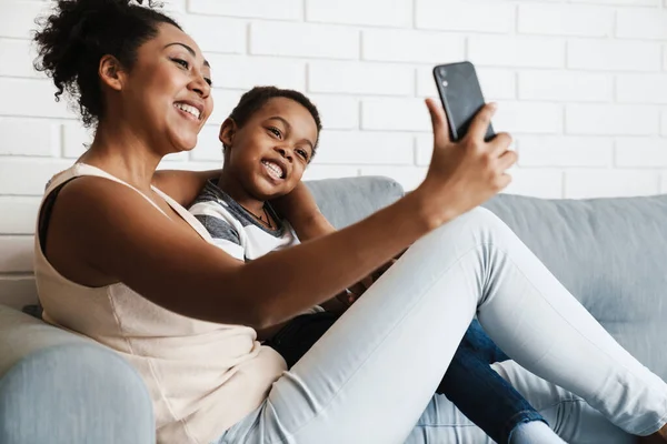 Black smiling woman taking selfie photo on cellphone while hugging her son at home