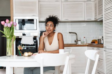 Evdeki mutfaktaki masada oturan siyahi kadın kağıtlarla çalışıyor.