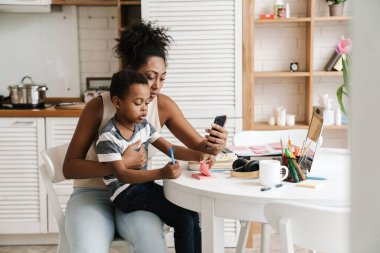 Siyahi anne ve oğul evde yan yana otururken yazıp cep telefonu kullanıyorlar.