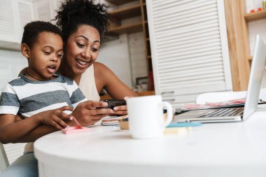 Siyah gülümseyen anne ve oğlu evde otururken cep telefonu ve laptop kullanıyorlar.