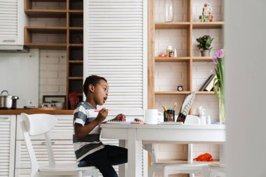 Siyah çocuk evde otururken dizüstü bilgisayar ve çizim yapıyor.