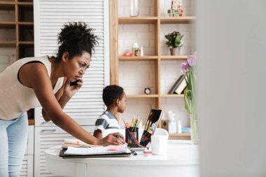 Evde dizüstü bilgisayarla çalışırken cep telefonuyla konuşan kafası karışık bir zenci kadın.