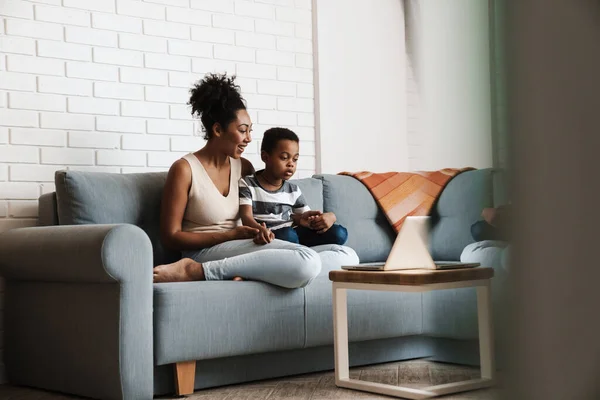 Black happy mother and son using laptop while sitting on sofa at home