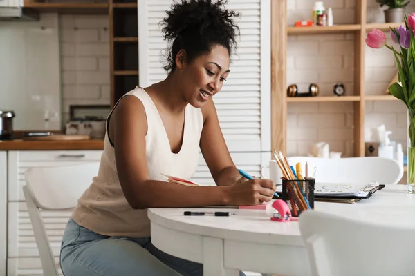 Black smiling woman working with papers while sitting at table in home kitchen