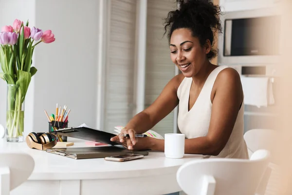 Black smiling woman using cellphone while working with papers at home kitchen
