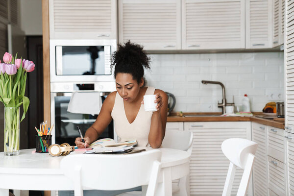 Black smiling woman drinking coffee while working at home kitchen