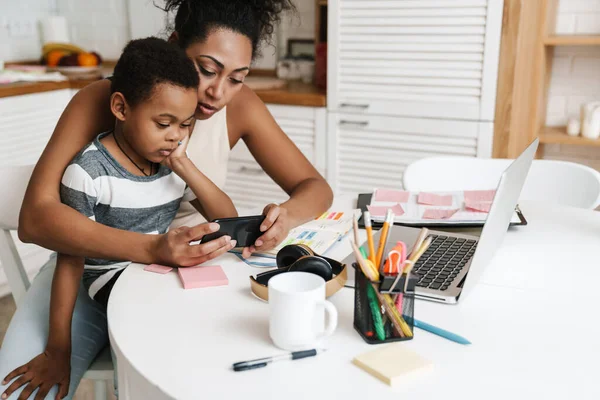 Black mother and son using mobile phone while sitting together at home