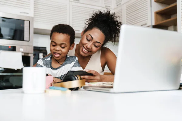 Black smiling mother and son using mobile phone and laptop while sitting together at home