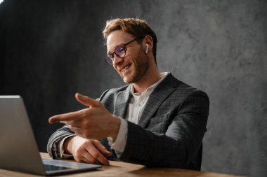 Ceketli ve gözlüklü gülümseyen adam ofiste masada otururken elleriyle işaret eden bir dizüstü bilgisayarla konuşuyor.