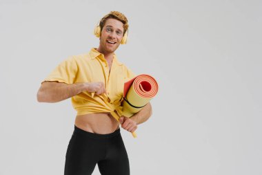 Gülümseyen beyaz eşcinsel adam spor paspası taşıyor. Kulaklıklı müzik dinliyor. Gri duvar arkasında duruyor.