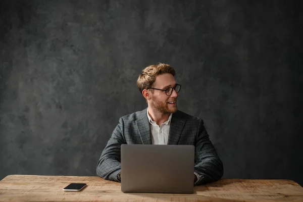 The laughing man in a jacket and shirt looking to the side while sitting in front of a laptop at a table in the studio