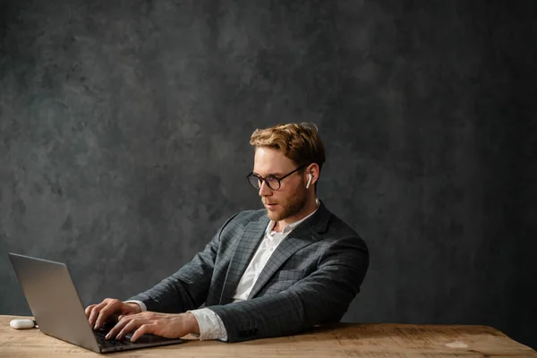 专心致志的戴眼镜和耳机的男人坐在工作室的桌子边 一边用笔记本电脑 — 图库照片