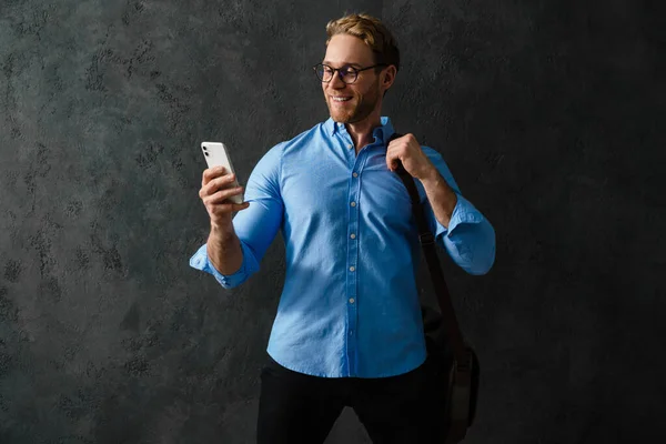 身穿蓝色衬衫 戴着眼镜 面带微笑的男子站在那里 肩上提着一个袋子 看着电话 — 图库照片