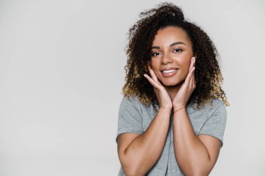 Gri stüdyoda dikilirken kameraya elleriyle bakan gülümseyen bir kadın.