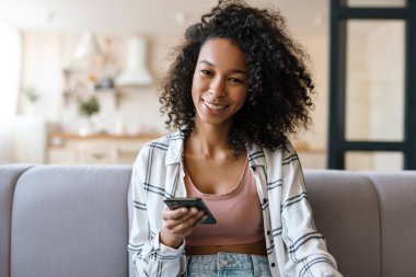 Sıradan giyinmiş gülümseyen Afrikalı genç bir kadın evdeki koltukta otururken cep telefonu takıyor.