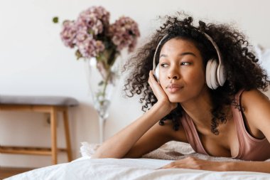 Orta yaşlı, Afrikalı bir kadın evde yatağında dinlenirken kulaklıkla müzik dinliyor, kapatıyor.
