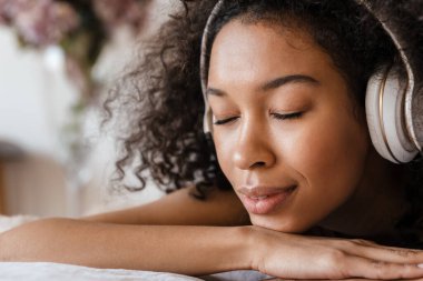Orta yaşlı, Afrikalı bir kadın evde yatağında dinlenirken kulaklıkla müzik dinliyor, kapatıyor.
