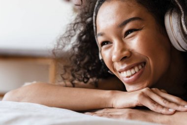 Orta yaşlı mutlu Afrikalı kadın evde yatakta dinlenirken kulaklıkla müzik dinliyor, kapat.