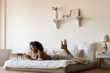 Orta yaşlı Afrikalı bir kadın evde yatağında dinlenirken kulaklıkla müzik dinliyor, elinde cep telefonuyla gülümsüyor.