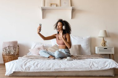 Orta yaşlı Afrikalı bir kadın evde yatağında dinlenirken kulaklıkla müzik dinliyor, selfie çekiyor ya da video çekiyor.