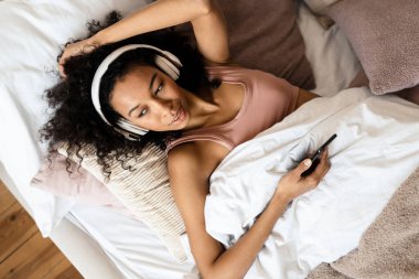 Yatakta cep telefonu tutan, kulaklıklı, gülümseyen Afrikalı genç bir kadının görüntüsü.