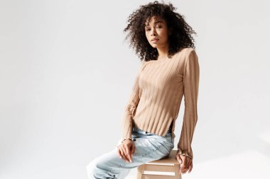 Genç Afrikalı kadın beyaz duvarın üzerinde izole bir şekilde otururken kameraya bakıyor.